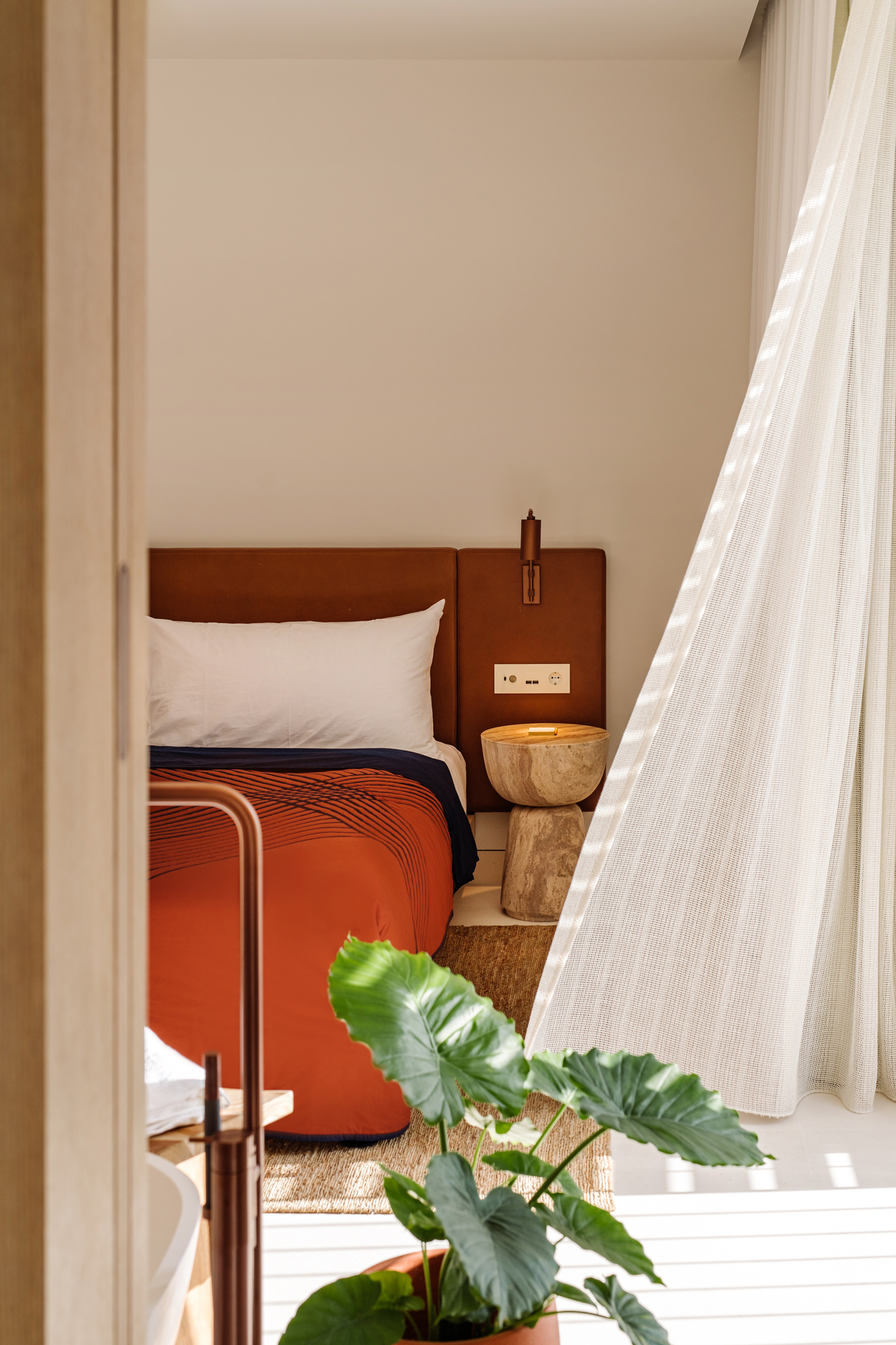 Schlafzimmer im The Standard Ibiza. Bett mit roter Bettwäsche. Link weht ein weißer Vorhang ins Bild. Ganz vorne steht eine grüne Pflanze.